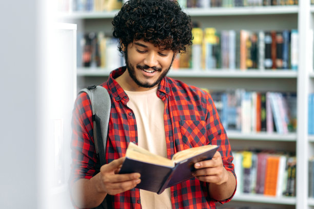 专注聪明积极的印度或阿拉伯大学或大学男生，站在图书馆的书架旁，读着有趣的书，准备考试，做作业，面带微笑图片下载