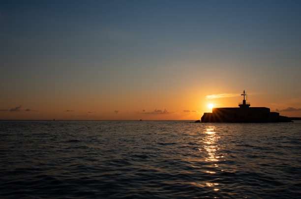 太阳在海上落下。天空闪烁着蓝色和橙色。光反射在波上。信号塔的影子在图片的一边，在太阳的前面。图片下载