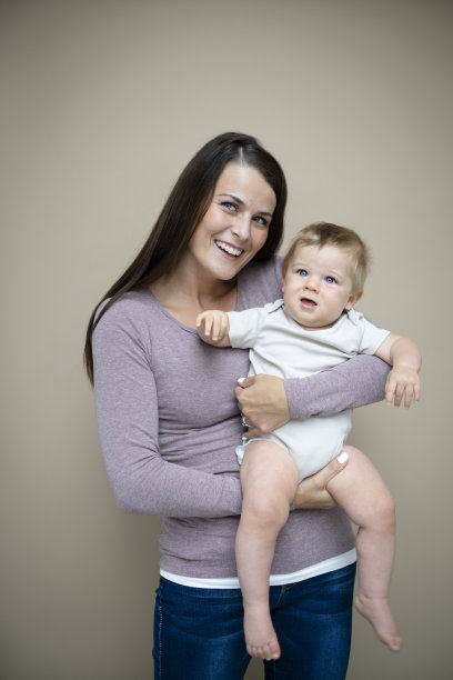 漂亮的年轻妈妈，深色头发，紫色上衣，抱着她7个月大的宝宝，站在棕色背景前图片下载