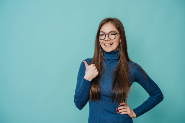 有吸引力的意大利年轻白种女人在蓝色毛衣和眼镜微笑广泛，显示大拇指向上的手势工作室的绿松石背景与空白的广告。模型，购物，促销，折扣。图片下载