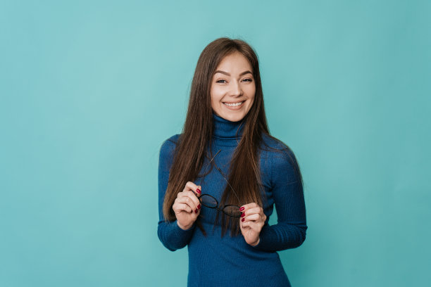 成功的年轻白人女性身穿蓝色毛衣，站在绿松石色的背景上，微笑着，拿着一副曾经戴过的眼镜。眼部矫正手术后不需要戴眼镜。模型、医药、成功图片下载