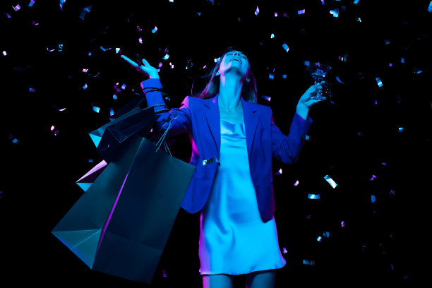 年轻时髦时髦的快乐女人与购物袋孤立在黑暗的背景在霓虹灯。购物，促销，广告，黑色星期五的概念图片下载