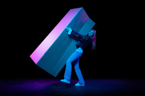 年轻兴奋的女人携带巨大的纸箱孤立在黑暗的背景霓虹灯。购物，促销，广告，黑色星期五的概念图片下载