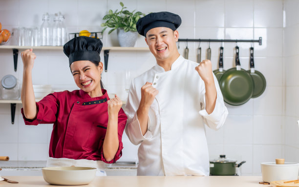 两名亚洲专业厨师夫妇穿着制服，戴着帽子，开心地微笑着，站着，摆着姿势，拿着厨房用具。餐厅、酒店、多元化概念。图片下载