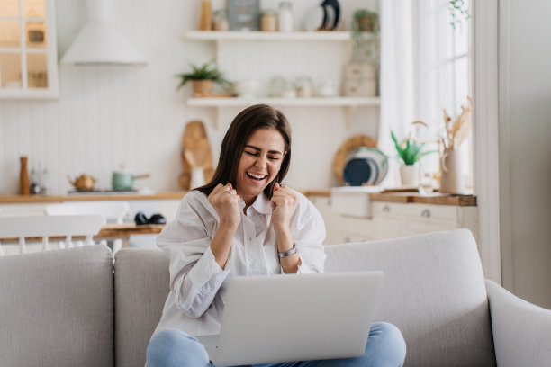 年轻的成功女性随意地坐在厨房的沙发上，拿着笔记本电脑握紧拳头，一副胜利者的表情，高兴地找到了一份新工作。女孩收到好消息，庆祝。快乐的人的概念图片下载