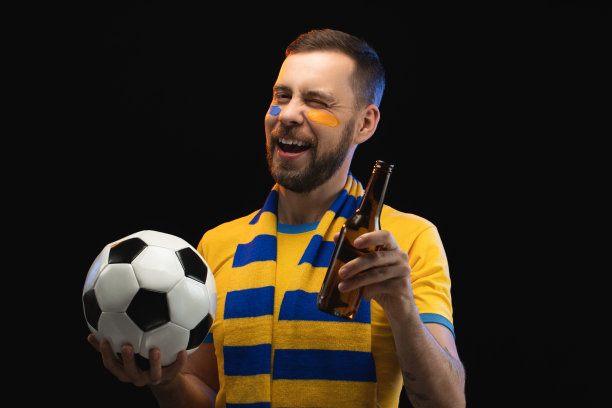 在摄影棚里，一名身穿黄蓝制服、笑容满面的球迷正在庆祝他最喜欢的球队获胜，他举着啤酒瓶，在黑色背景上拿着一个球摆姿势，眼睛眨巴着。图片下载