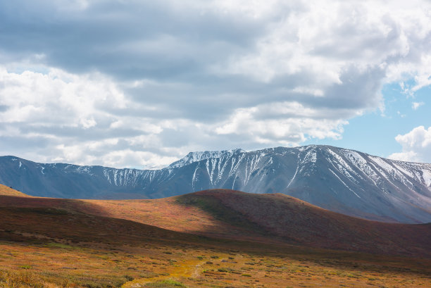 秋天，高原上的山丘和阳光照耀下的雪山在戏剧性的多云天空下。山中鲜艳的秋色。天气多变时的阳光和乌云。图片下载