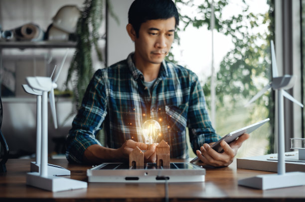 一位工程师坐在那里，拿着一个灯泡和一台平板电脑，看着一座房子的模型。桌子上有太阳能电池板和房子模型。设计利用风能和太阳能等可再生能源。图片下载