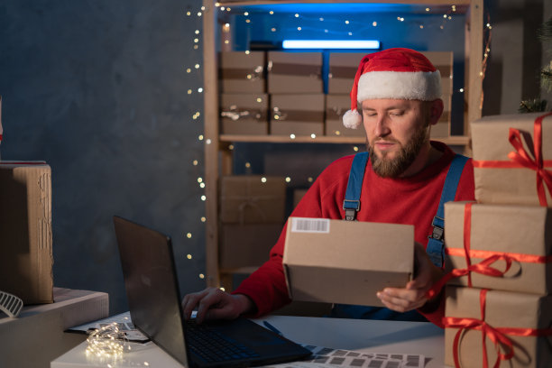 圣诞老人戴着帽子，拿着礼品盒，用笔记本电脑坐在家里的办公桌旁。电子商务网站圣诞假期网上购物电子商务销售图片下载