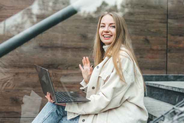 视频通话的概念。年轻的白人金发女学生坐在楼梯上微笑着对着屏幕挥手图片下载