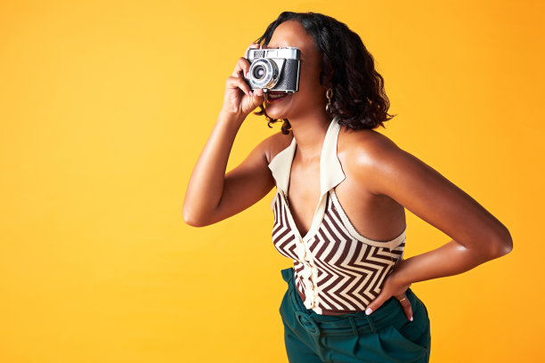 相机，摄影师和富有创造力的女人在摄影工作室的背景墙。快乐的黑人女孩和模特在黄色的背景下拍摄设计师时装的数码照片图片下载