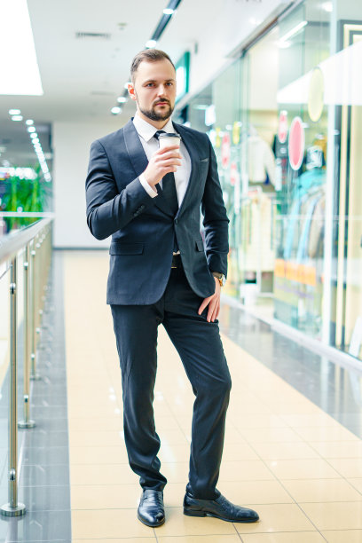 一个留着胡子的年轻男子，穿着西装，拿着一杯咖啡在商场里。图片下载