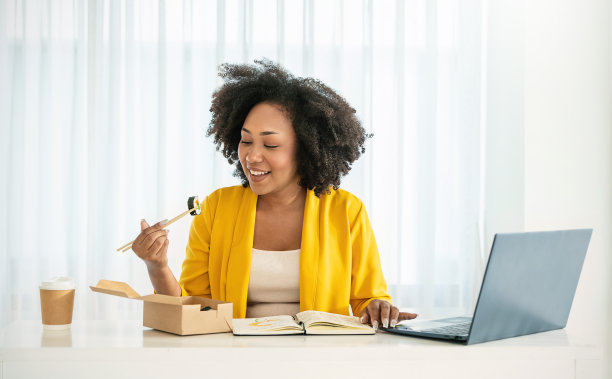 年轻的非洲女性享受中餐在办公室午休，女员工拿着筷子吃外卖日本或韩国寿司午餐盒饭在工作场所在家庭办公室的肖像。图片下载