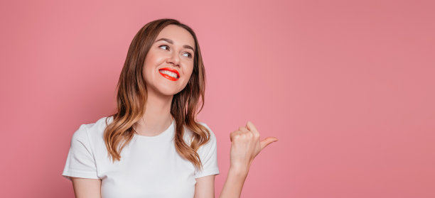 快乐微笑的年轻女子在白色t恤指着拷贝空间和看远离孤立的粉红色工作室背景。网页横幅。副本的空间。图片下载