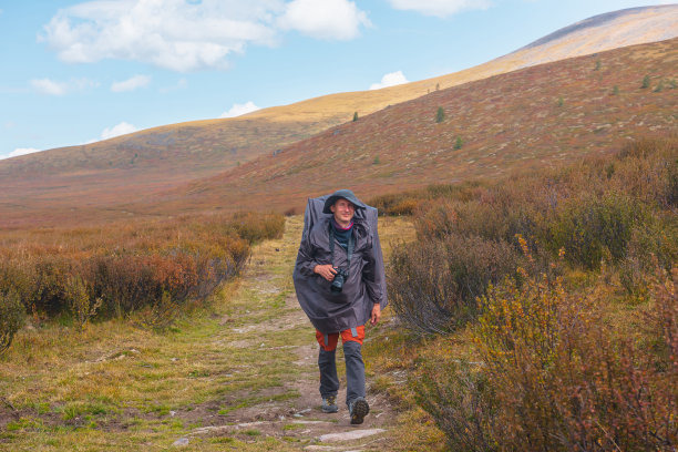 一个人穿着雨衣，背着大背包，走在高山高原渐隐的秋草丛中。雨天登山时带着相机的背包客。图片下载