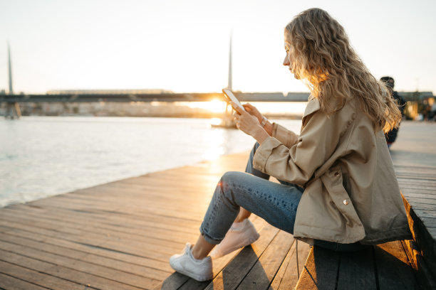 穿着大衣的年轻休闲女子坐在海堤上的长凳上使用智能手机图片下载