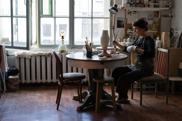 车间里，身穿围裙的年轻女子坐在圆桌旁的椅子上，为手工制作的陶瓷花瓶作画图片下载