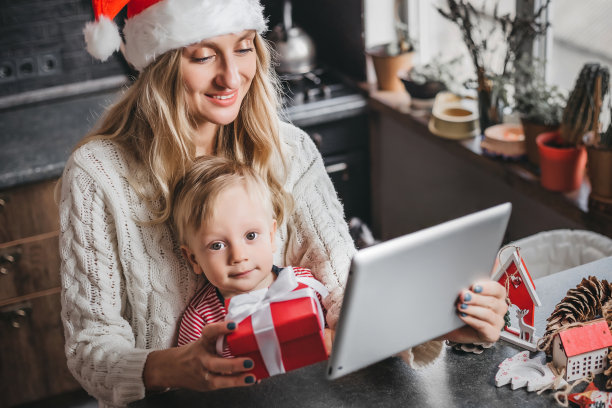 戴着新年帽、穿着白色毛衣的母亲给儿子看平板电脑上的游戏，陪他度过闲暇时光。妈妈在网上商店为她的儿子订购玩具图片下载