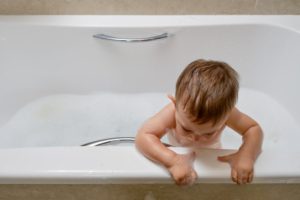 蹒跚学步的婴儿爬过浴缸边缘，冒着摔倒的危险。把孩子一个人留在浴室里的危险图片下载