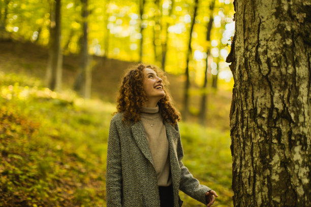 秋天的森林里，穿着大衣的红发卷发美丽女孩。享受假期的漂亮女人。参观当地的旅游景点。女孩在自然中笑。感觉幸福。迷人的微笑。快乐的女士图片下载