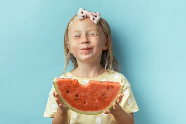 可爱的小女孩在蓝色的背景下享受着一大片鲜红多汁的西瓜。穿着黄色t恤的金发快乐白人女孩的肖像。炎热的夏天的概念图片下载