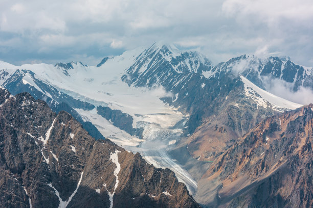 在多云的天空下，有着长长的冰舌和高高的雪山。美丽的鸟瞰图，阳光照耀下的大冰川和云中雪山。俯瞰雄伟的山脉。图片下载