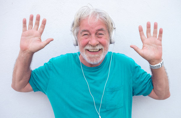 穿着蓝色t恤，戴着耳机，这位快乐的老人低头看着镜头，举起手投降——他是一位迷人的大胡子快乐老人图片下载