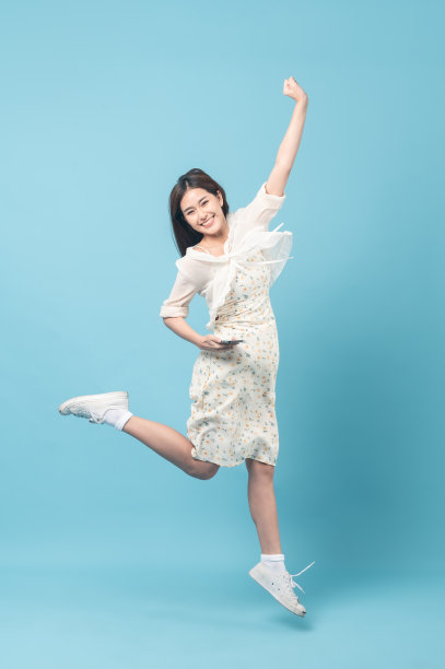 美丽的年轻亚洲女人在白色连衣裙花图案使用智能手机和跳跃隔离在蓝色背景图片下载