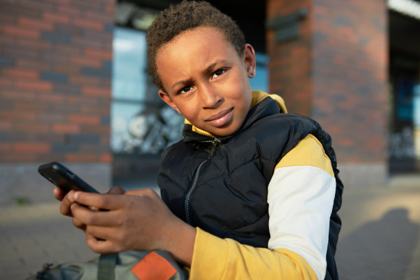 皮肤黝黑的困惑男孩使用智能手机，试图了解如何使用新的应用程序，坐在附近的城市街道的人行道上，看着摄像头严肃的脸图片下载