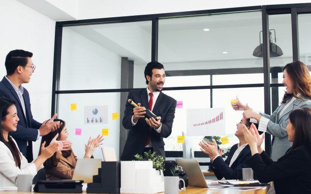 白人帅老板拿着一瓶酒，向办公室的亚洲团队表示祝贺。团队合作，业务和工作理念。图片下载