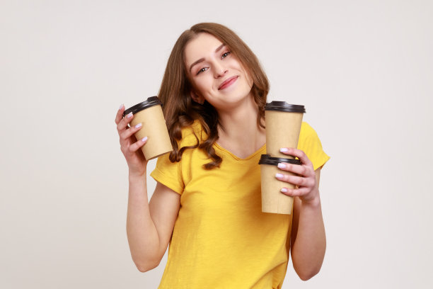 送货服务。友好快乐的少女年轻的年龄在黄色t恤拿着咖啡，饮料在一次性杯子，看着相机微笑。图片下载