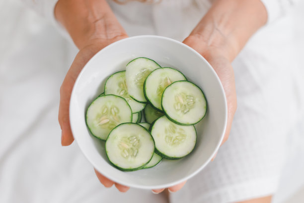年轻的白种人妇女拿着一碗黄瓜片准备在家里的美容护肤日常。迷人的快乐女士与裸露的脸在浴袍做皮肤治疗。图片下载