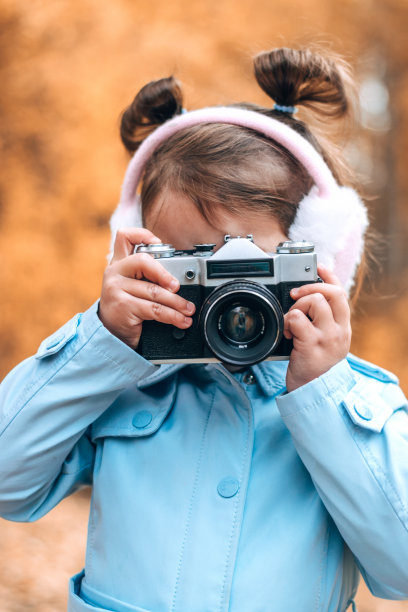 可爱的白人小女孩穿着蓝色雨衣和毛皮耳机在秋天的公园里用相机拍照。秋天的概念图片下载
