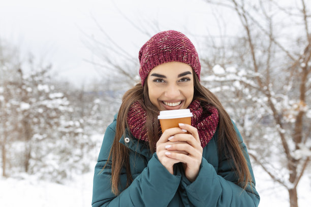 漂亮的年轻女子在冬季装备持有一次性杯子装满热咖啡或茶。女孩手捧热饮，在冬天走在户外。图片下载