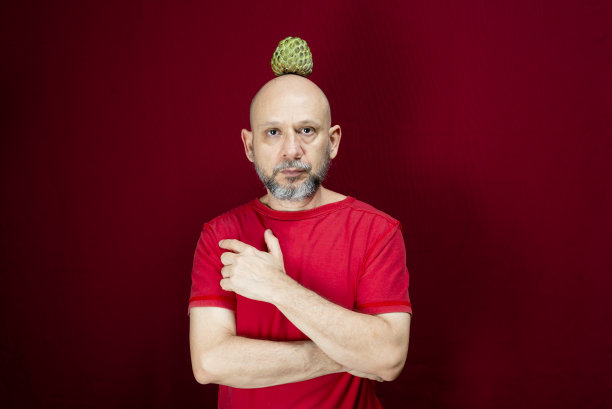 年轻英俊的男子与胡子，光头和红衬衫站与松果平衡的头在红色的背景。图片下载
