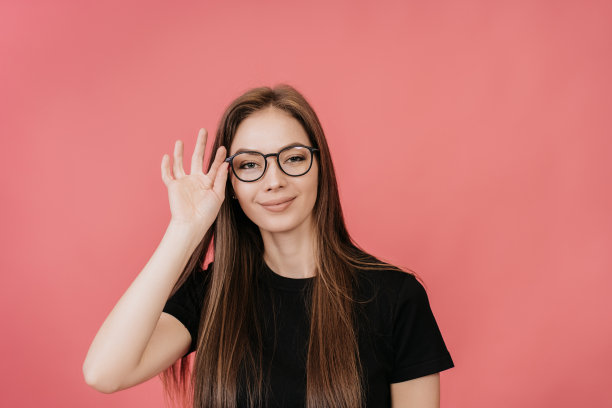可爱的金发女郎戴着眼镜，穿着黑色t恤，一头华丽的长发，站在粉红色背景的工作室里，调整了一下眼镜，看着镜头，微笑着。图片下载