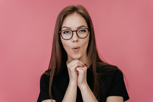 一位戴着眼镜、身穿黑色t恤的美丽意大利女子在身前交叉双手，惊讶地在演播室的粉色背景上发出声音。惊讶的女模特疑惑。销售,折扣。图片下载