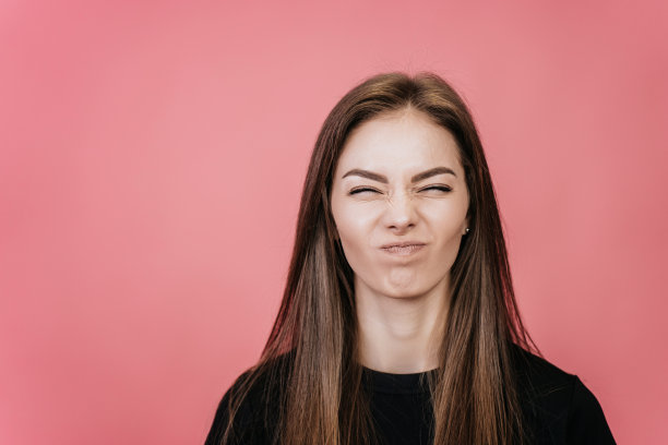 在一张特写照片中，一个不满的女孩，留着长发，穿着黑色t恤，在粉红色的背景下，满脸皱纹，闭上了眼睛。带有空背景的布局。被激怒的模特在工作室图片下载