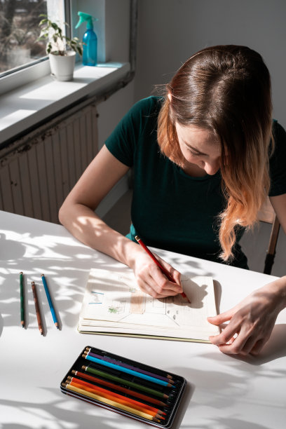 穿着绿色t恤的年轻女子坐在靠窗的桌子旁，用铅笔画画。图片下载