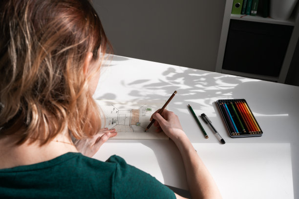 穿着绿色t恤的年轻女子坐在桌子旁用铅笔画画。图片下载