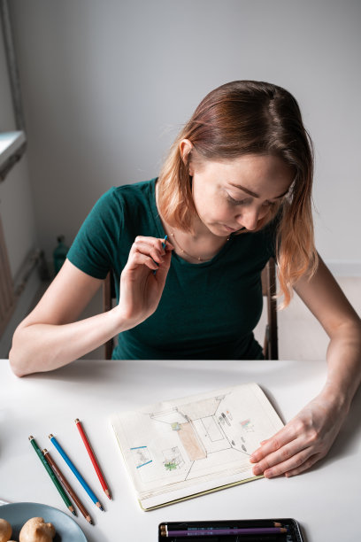 穿着绿色t恤的年轻女子坐在靠窗的桌子旁，用铅笔画画。图片下载