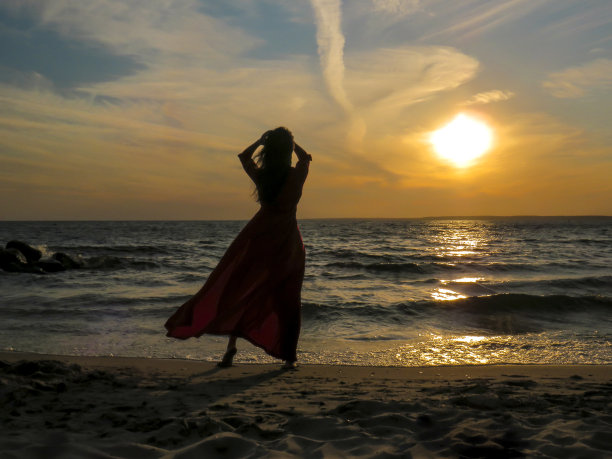 一个迷人的女人在海边穿着红色的裙子，背景是日落时的波浪图片下载