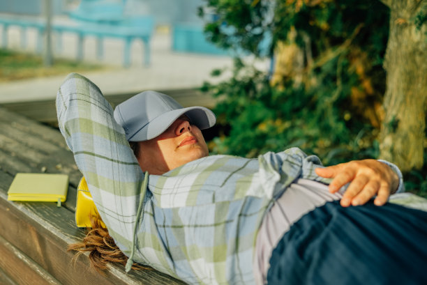 夏天，一个年轻的女子躺在一张木凳上，用棒球帽遮住了她的脸图片下载