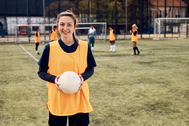 快乐的女足球运动员在球场上拿着球，看着镜头。图片下载