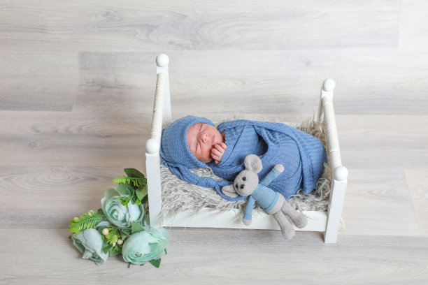 裹着蓝色毛巾、头部包裹着的新生男婴，躺在小床上，周围有花朵和树叶图片下载