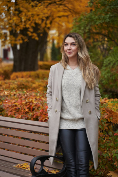 秋日公园里的美女，身穿淡大衣，站在长椅旁。黄树和落叶。图片下载