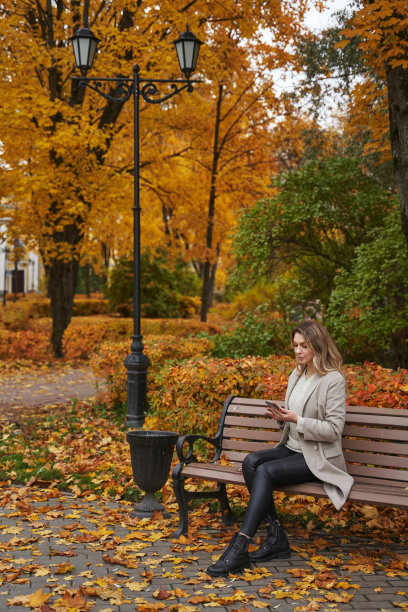 在公园里，一位美丽的女子以秋天的树木为背景，坐在长椅上，看着智能手机里的东西。以落叶为背景的美丽的秋天肖像图片下载