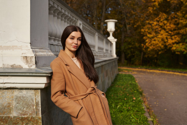 秋天的公园里，一位身穿棕色大衣的美女微笑着倚着白色的柱子站着图片下载