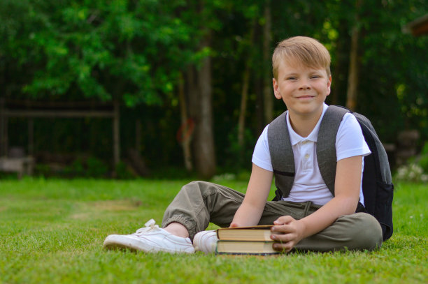 一个带着公文包微笑的白人男孩坐在草坪上，读着一本书，这是关于教育的概念。图片下载