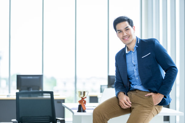 高兴的亚洲年轻商人看到一个手腕成功的穿着西装的男人，穿着蓝色夹克，在办公室的桌子上的冠军杯。图片下载